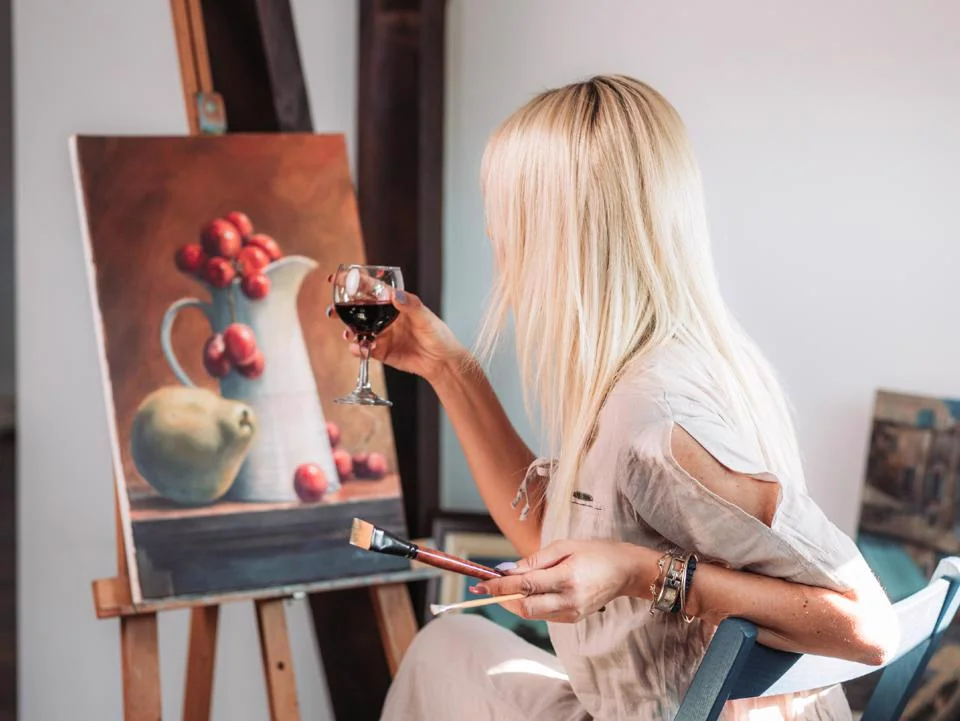 Woman painting at an easel while enjoying a glass of red wine